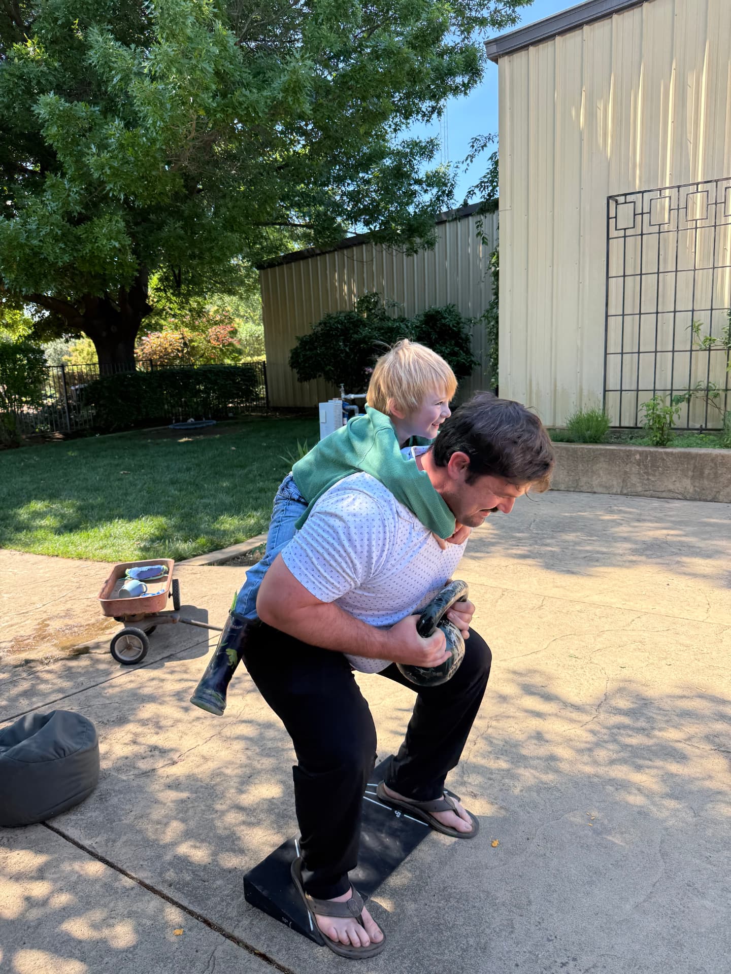 A father does a kettlebell squat while his young son rides on his shoulders during a beginner fitness comeback workout at home in Chico, CA, showing the family-friendly approach of mobile personal training with Gym Truck Chico.