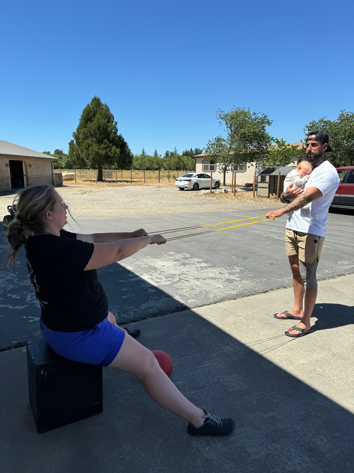 A mother completes a resistance-band row while a personal trainer holds her sleeping baby, showing how beginner fitness comebacks work with family-friendly, mobile personal training in Chico, CA from Gym Truck Chico.