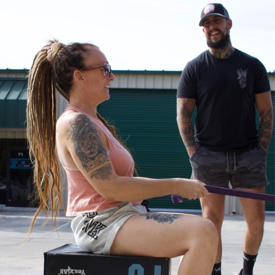 A certified NASM personal trainer coaches a woman through a seated resistance-band row outside on a driveway in Chico, CA, demonstrating expert at-home fitness instruction from Gym Truck Chico.