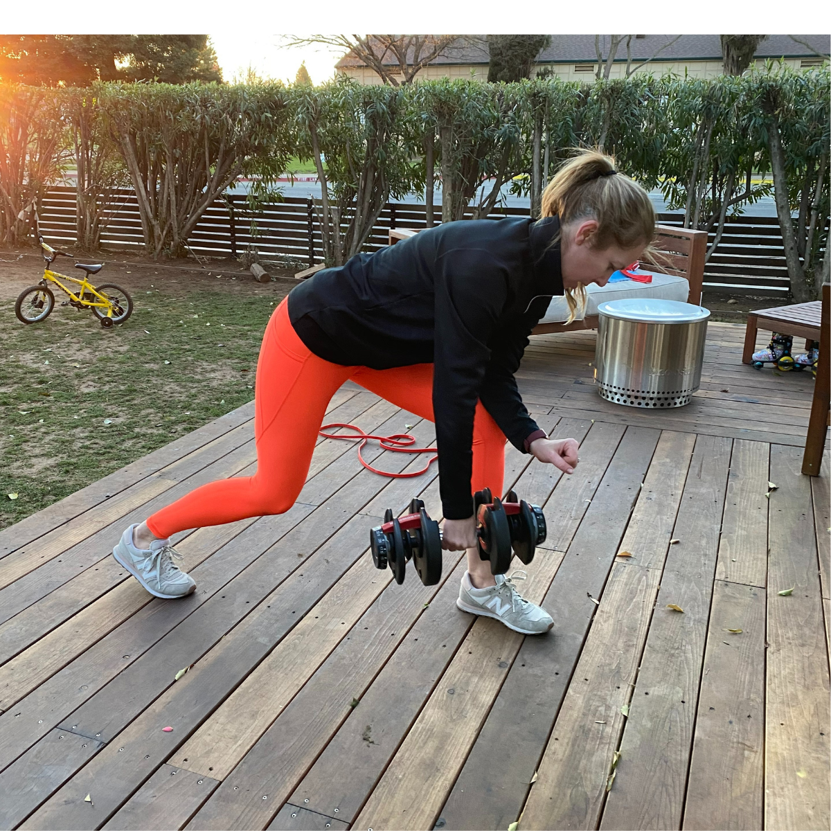 A woman in bright orange leggings performs a controlled dumbbell row on a residential wooden deck during a private, fully customized women's fitness session with Gym Truck Chico in Chico, CA.