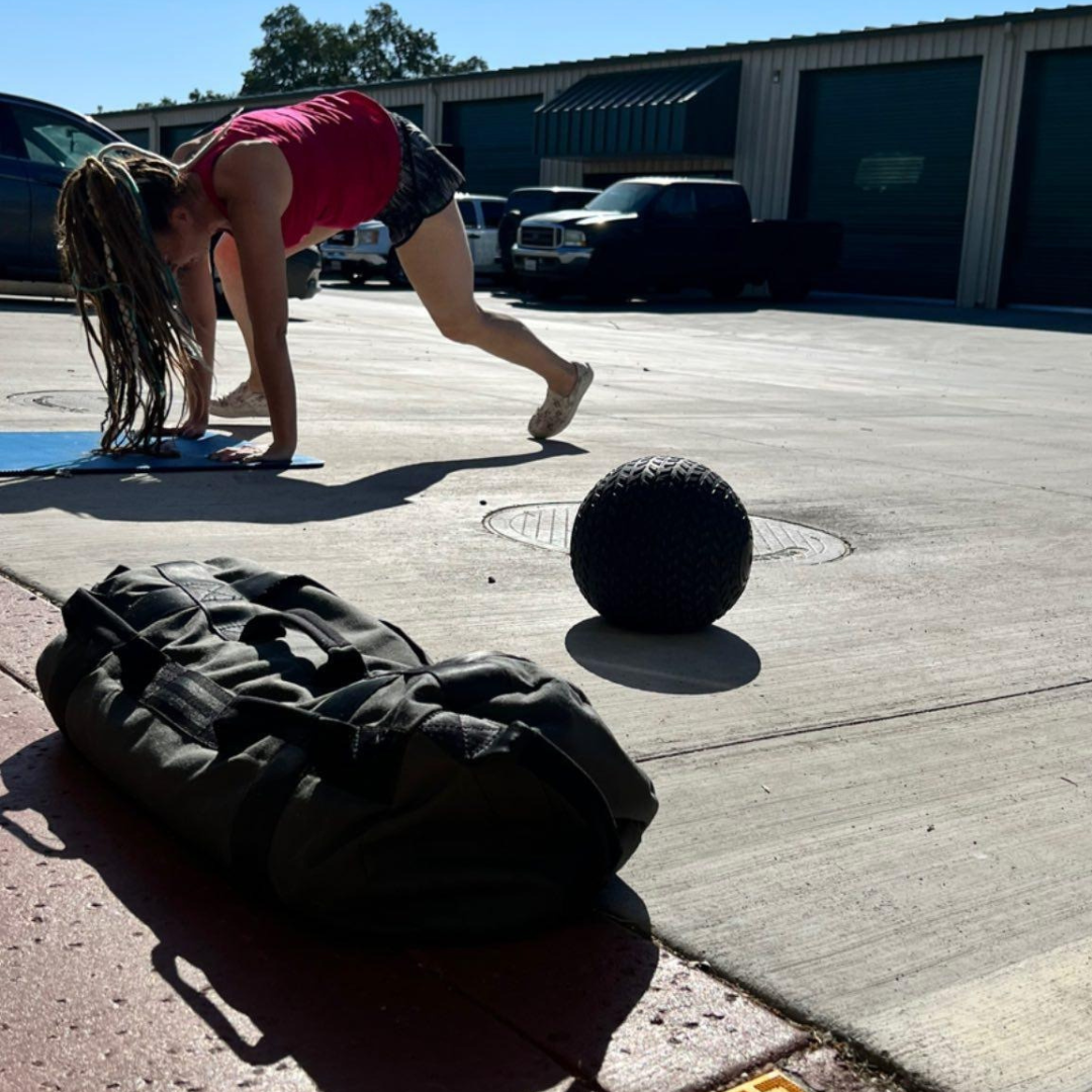 A woman performs a hip and knee mobility and flexibility workout on a yoga mat in an outdoor Chico, CA driveway, with a slam ball and gym bag nearby, showing the joint-health focus of mobile at-home personal training with Gym Truck Chico.