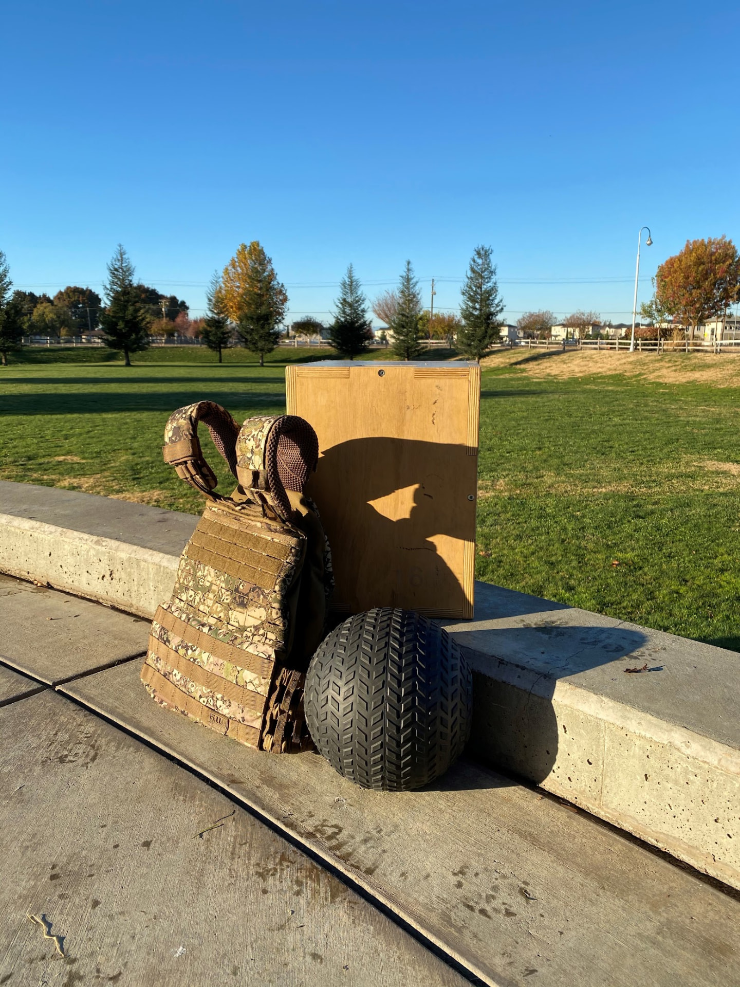Camouflage weighted training vest, a wooden plyo box, and a rugged tire arranged on a concrete ledge at a sunny Chico, CA neighborhood park, showing how Gym Truck Chico brings convenient, fully customized outdoor workouts to local parks and driveways.