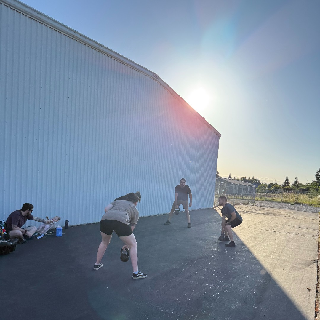A small group of adults performs kettlebell training together outdoors on a sunny morning beside a white building, with a coach nearby and a parent watching a baby, showing the customized small-group personal training experience of Gym Truck Chico in Chico, CA.