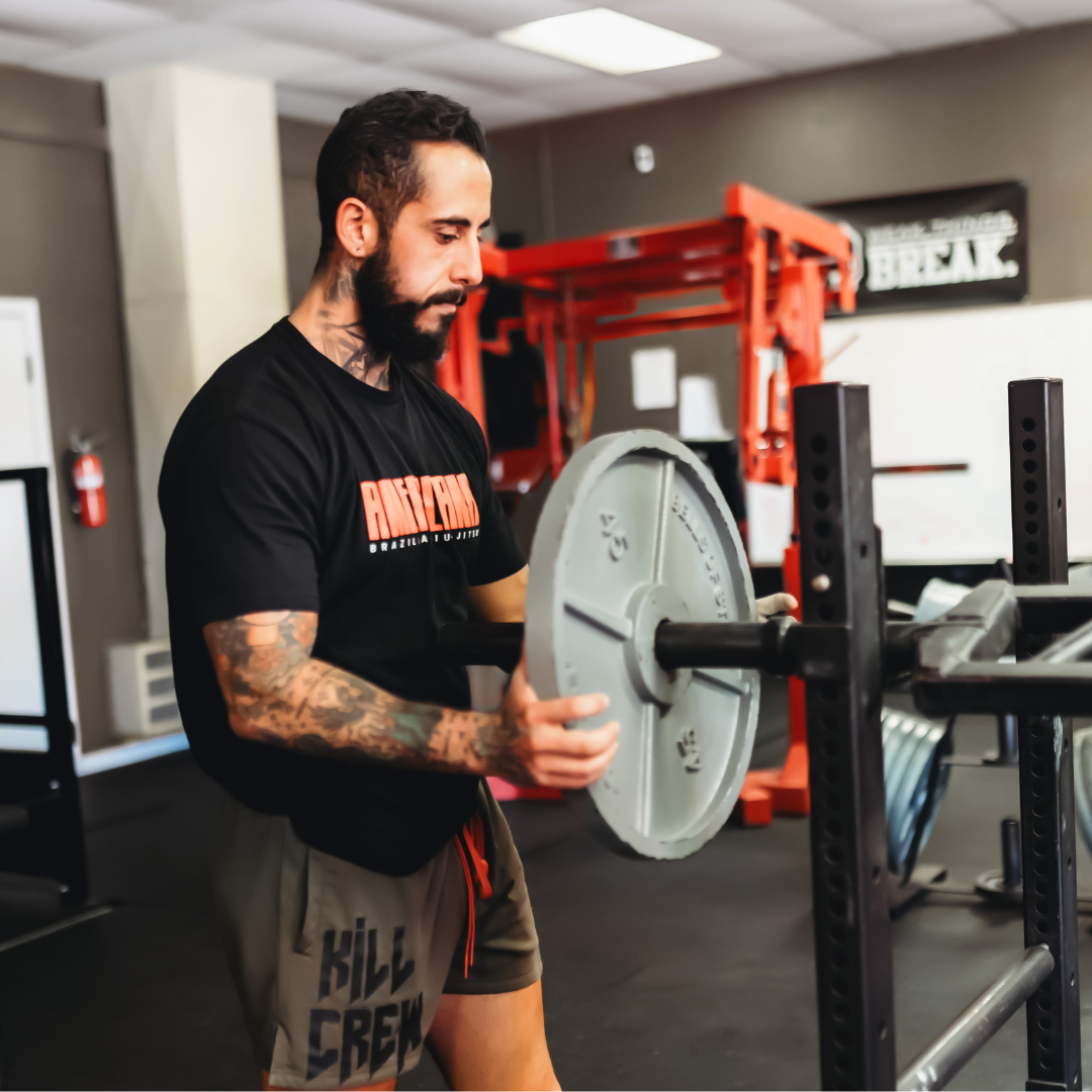 A certified personal trainer loads an iron weight plate onto a barbell in a private gym setting, demonstrating the progressive machine-supported strength training behind fully customized muscle-building and hypertrophy sessions with Gym Truck Chico in Chico, CA.